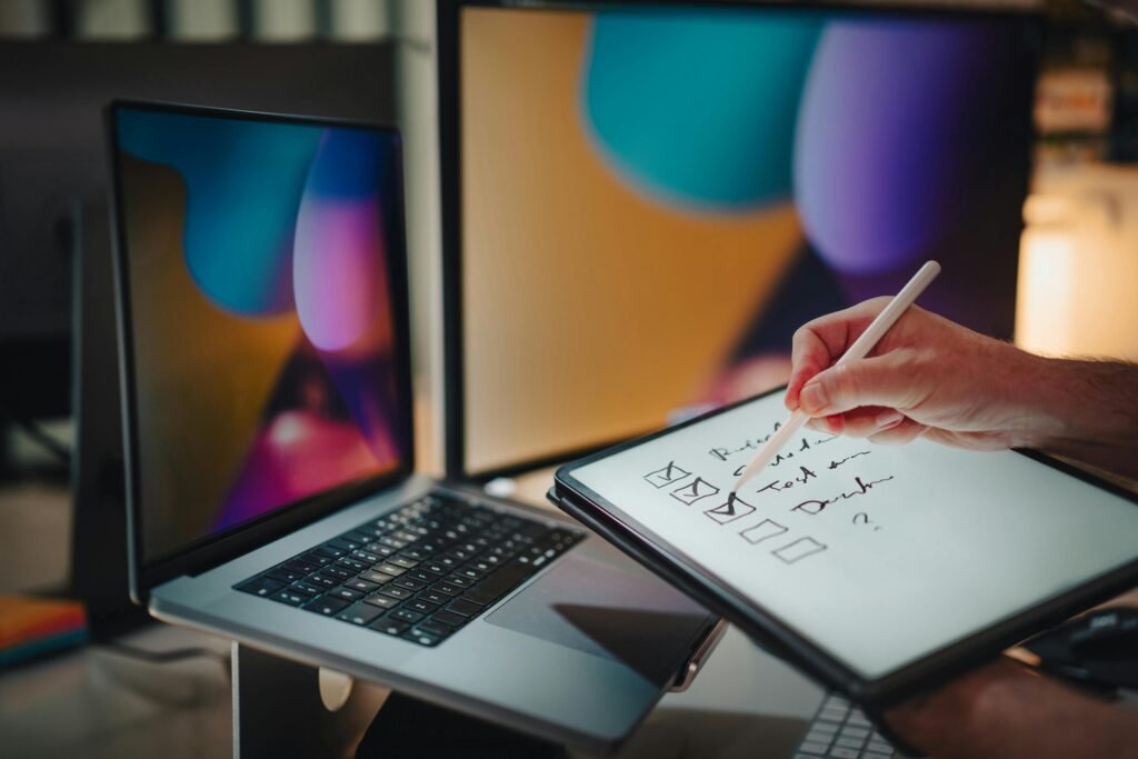 an individual writing on ipad with electronic pen and open window laptop